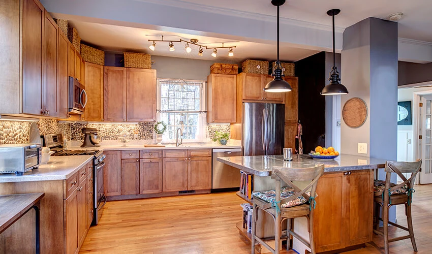 Modern kitchen with wooden cabinets, stainless steel appliances, and a central island with bar stools, illuminated by pendant and track lighting.